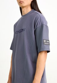 Camiseta de algodón gris de manga corta con cuello redondo, que presenta un sutil gráfico de automóvil en el frente y un logo de BMW Motorsport en la manga.