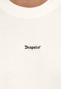 Bijela pametna majica s kratkim rukavima i okruglim izrezom, na kojoj je sa prednje strane prišiven pravokutni crni simbol s natpisom "Dropsize". Mekana tekstura, minimalistički dizajn.