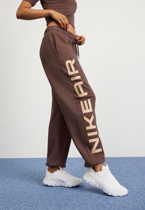 Personne portant un pantalon de survêtement Nike Air marron avec un grand logo beige sur le côté et des baskets blanches, debout sur un sol recouvert d'un tapis bleu.