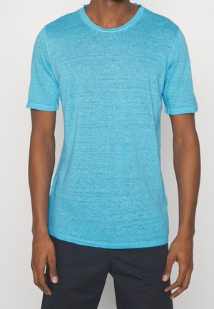Mand iført en lyseblå t-shirt med korte ærmer og mørke bukser, stående mod en ensfarvet baggrund.