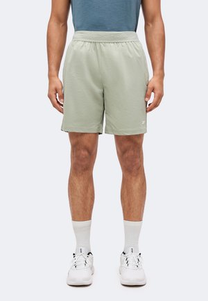 Homme portant un short de sport vert clair, des chaussettes blanches et des baskets blanches Reebok, debout devant un fond uni.