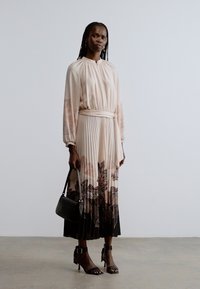 Lang plisseret kjole i blød lyserød med blomsterprint, spændt ind i taljen, kombineret med sorte remhæle og en lille sort håndtaske.