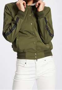 Olivgrön bomberjacka med dragkedja och tryckknappar, ribbade ärmändar och kontrasterande svarta ränder på ärmarna. Matchas med vita byxor.