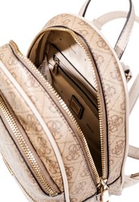 Mochila beige con un exterior texturizado y con un patrón, y cremalleras doradas. El interior cuenta con múltiples bolsillos y una etiqueta de la marca, fabricada con materiales duraderos.