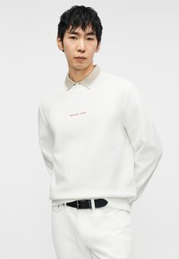 Λευκό φούτερ με μπεζ γιακά, που φέρει κόκκινο λογότυπο «Michael Kors». Συνδυάζεται με λευκό παντελόνι και μαύρη ζώνη.