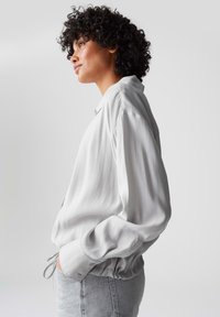 Helle graue Satinbluse mit lässiger Passform, einem Button-Down-Kragen und elastischen Bündchen. Der Stoff hat eine glatte, glänzende Textur.