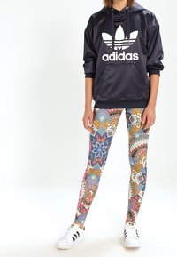 Svart huvtröja med vitt Adidas-logga, ihop med färgglada mönstrade leggings med blommiga och geometriska mönster. Vita sneakers.