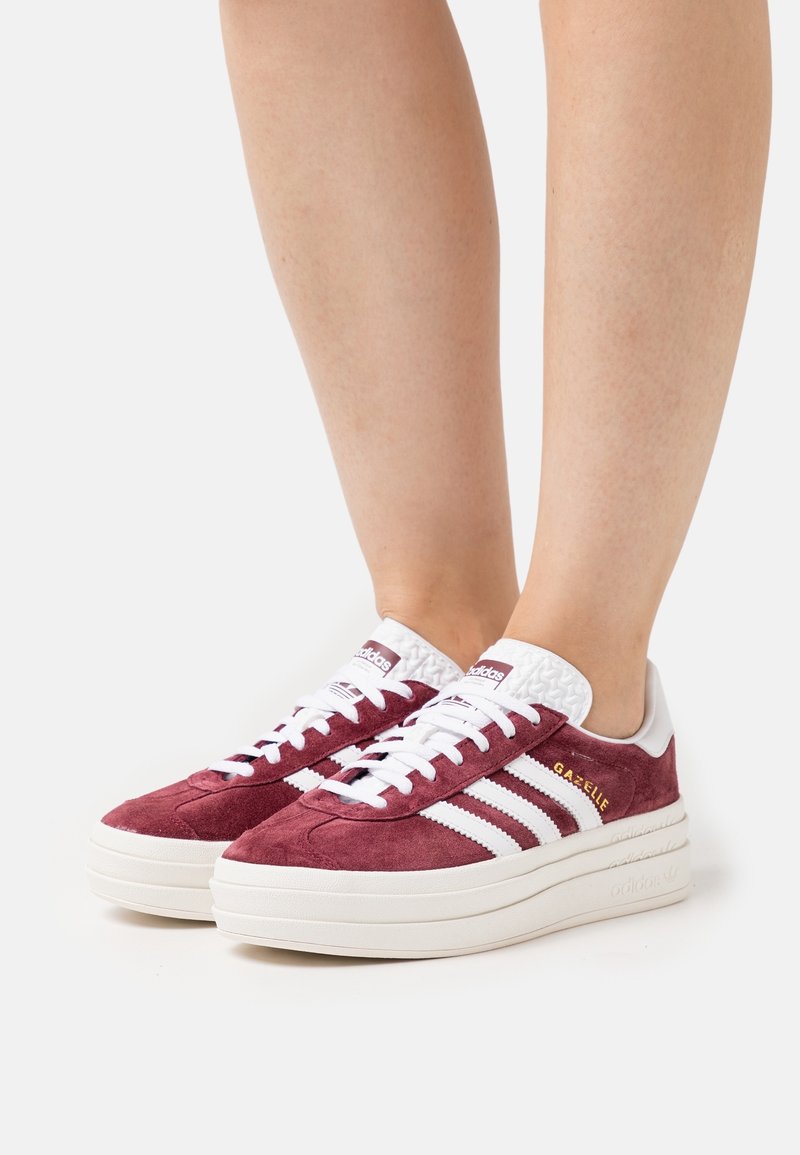 Henkilö, joka on pukeutunut viininpunaisiin Adidas Gazelle -tennareihin, joissa on valkoiset raidat ja paksut valkoiset pohjat, seisoo valkoisella pinnalla.
