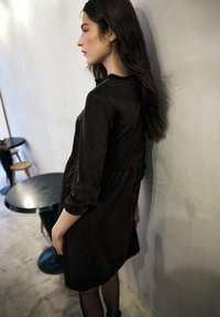 Schwarzes Wildlederkleid mit langen Ärmeln und geraffter Taille. Das Modell steht neben einer grauen Wand, im Hintergrund schwarze Möbel.