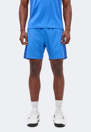 Homme portant un short et un t-shirt de sport bleus, des chaussettes blanches et des baskets Adidas blanches, debout devant un fond clair uni.