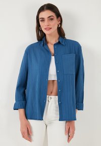 Blaues strukturiertes Hemd mit Knopfverschluss und Brusttasche, lange Ärmel, die an den Manschetten hochgerollt sind, über einem weißen Top und weißen Hosen getragen.