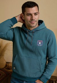 Turquise gestreepte hoodie met een voorkeurszak, een capuchon met trekkoord en een patch met een geometrisch ontwerp en tekst. Zachte textuur, casual pasvorm.