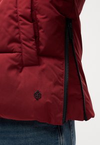 Chaqueta de plumas marrón con tejido texturizado, cremallera negra y un pequeño parche negro con el logo cerca del dobladillo. Detalle de costura sutil presente.