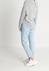 Helle graue Strickjacke mit lockerer Passform; kombiniert mit hellblauen Jeans mit Schnürdetails an den Bündchen und weißen Sneakers.