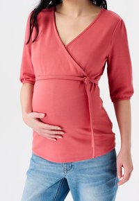 Top corallo avvolgente con scollatura a V, maniche 3/4 e vita regolabile con laccetto, abbinato a jeans azzurri chiari. Tessuto morbido e elastico visibile.
