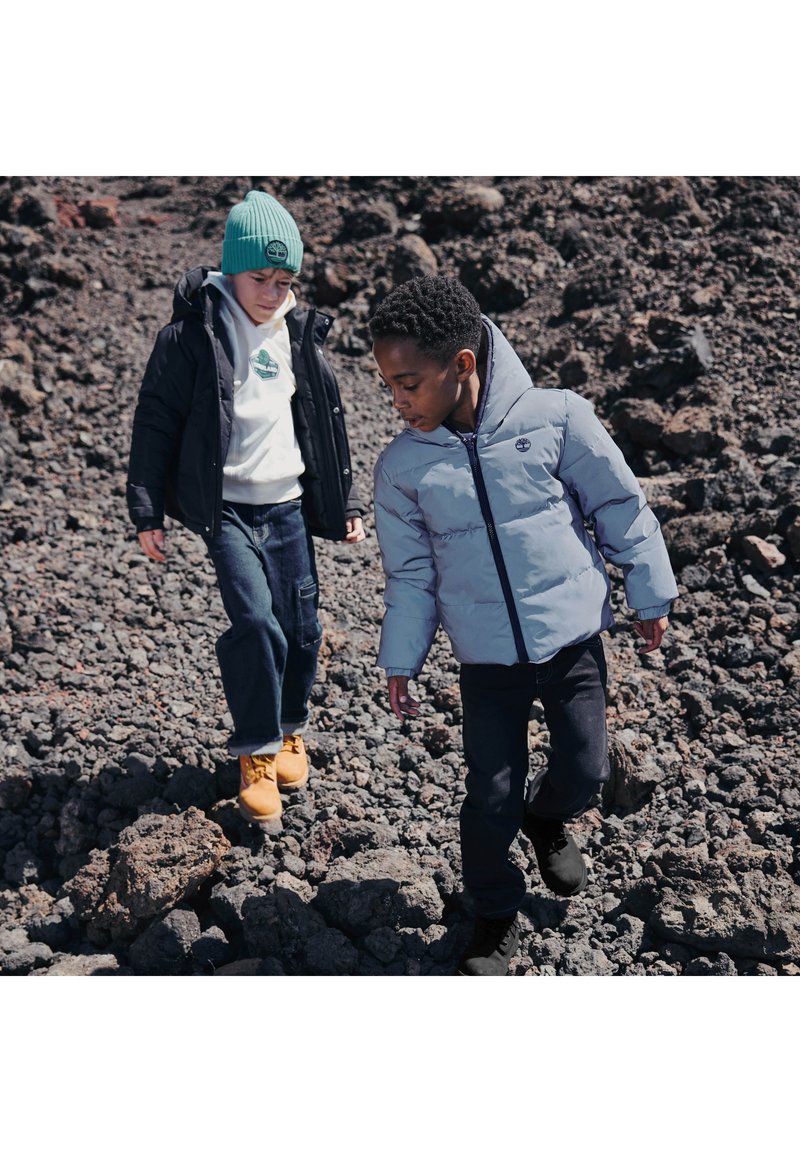 Deux enfants marchent sur un terrain rocheux. L'un porte une doudoune gris clair et un pantalon noir, tandis que l'autre a un manteau noir, un bonnet vert et un jean bleu.