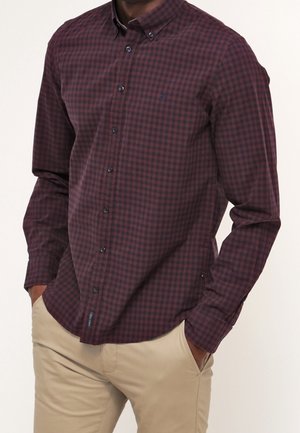 Homme portant une chemise à manches longues à carreaux bordeaux et noirs avec un pantalon beige, les mains dans les poches, devant un fond uni.