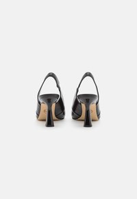 Tacones slingback de piel negra con un tacón curvado, acabado suave y suela beige. El diseño enfatiza formas elegantes y estilizadas.