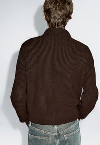 Mand iført en mørkebrun ribbet striktrøje og falmede blå jeans, vist fra ryggen mod en ensfarvet lys baggrund.