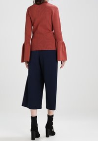 Rostfarbener, gerippter Pullover mit ausladenden Ärmeln, kombiniert mit navyblauen, weiten Culottes und schwarzen Ankle-Boots mit goldenen Knopfakzenten.
