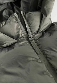 Giacca puffer con un esterno di un verde oliva opaco, una cerniera robusta e un marchio sottile lungo il nastro della cerniera; leggera e con una texture liscia.