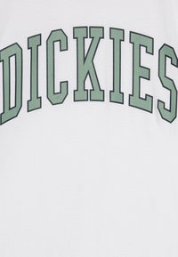 Balta marškinėlių su ryškiai žaliais užrašais, kurie rašo "DICKIES" išlenktoje formoje. Medžiaga atrodo minkšta ir vientisa.