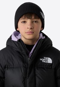 Svart dunjakke med glidelås foran og høy krage, med en grå logo. Brukes over en lilla hettegenser, toppet med en ribbestrikket svart lue.