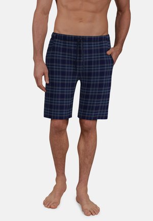 Male torso and legs wearing navy blue plaid drawstring shorts, one hand in pocket, standing barefoot against a plain background.