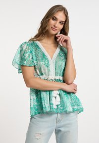 Blusa verde a fantasia con maniche corte a sbuffo, scollo a V, bordo in pizzo e nappine bianche in vita, abbinata a jeans chiari strappati.