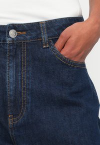 Main reposée à l'intérieur de la poche avant d'un jean en denim bleu foncé avec des coutures visibles et un bouton en métal.