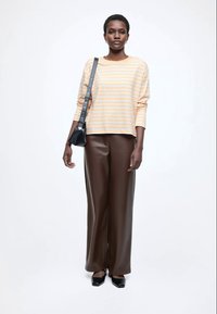 Bluzka w paski z długim rękawem w kolorze brzoskwiniowym i białym, zestawiona z wysokim stanem brązowych skórzanych spodni. Modelka trzyma małą czarną torbę.