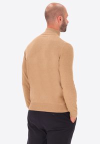 Maglione beige a collo alto realizzato in tessuto morbido, design aderente, orlo a costine, texture liscia e stile minimalista, indossato con pantaloni scuri.