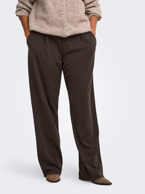 Person iført løstsiddende mørkebrune bukser, beige, fluffy, knappet sweater og brune spidse sko, stående med hænderne i lommerne mod en ensfarvet baggrund.