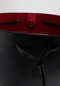 Sac seau en cuir noir avec un intérieur rouge contrastant, doté d'une fermeture à cordon et d'une quincaillerie métallique décorative.