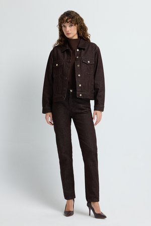 Dunkelbraune Jeansjacke mit Fronttaschen, bronzefarbenen Knöpfen und passenden, enganliegenden dunkelbraunen Hosen. Das Model trägt High Heels vor einem hellen Hintergrund.