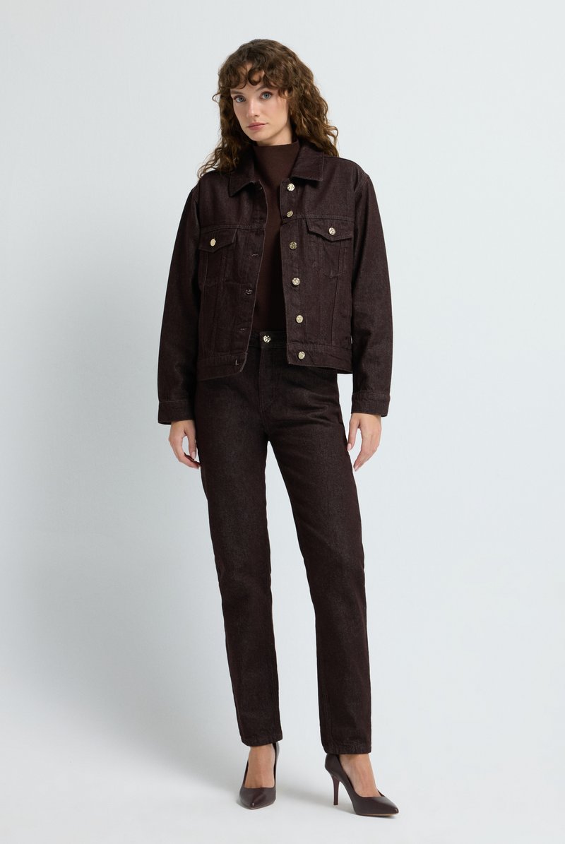 Chaqueta de mezclilla marrón oscuro con bolsillos frontales, botones de bronce y pantalones ajustados a juego en marrón oscuro. La modelo lleva tacones, contra un fondo claro.
