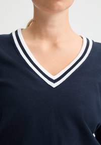 Marineblauwe V-hals trui met witte en donkerblauwe gestreepte ribbelkraag. Zachte textuur met een getailleerd ontwerp dat de halslijn accentueert.