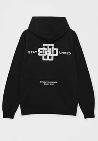 Sweat à capuche noir avec logo stylisé blanc au centre indiquant "STAY UNITED" et texte plus petit en dessous mentionnant "STWD TRADEMARK DEPUIS 2018".