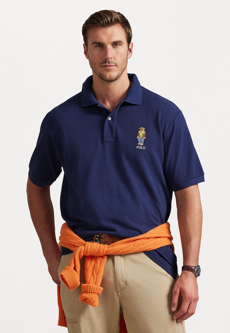 Tumši zils polo krekls ar teksturētu audumu un īsām piedurknēm, ar izšūtu lāča logotipu un "POLO" tekstu. Oranža džemperis sasietā veidā ap vidukli.