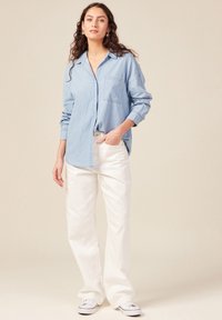 Camisa de chambray de manga larga en azul claro con un bolsillo en el pecho, combinada con pantalones anchos blancos y zapatillas blancas sobre un fondo neutral.