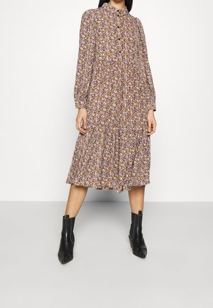 Femme portant une robe fleurie à manches longues, longueur genou, associée à des bottines noires, debout devant un fond blanc uni.