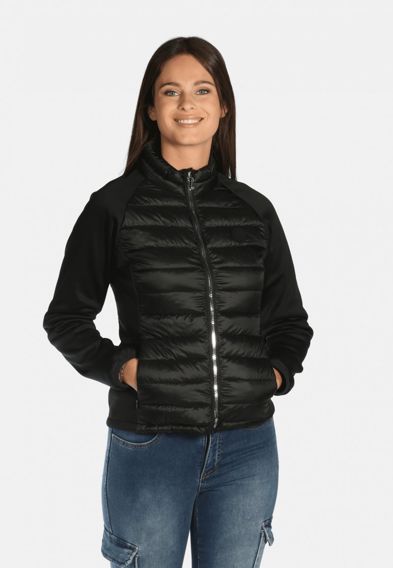 Veste noire avec un devant matelassé et rembourré, manches noires ajustées, fermeture éclair, col montant et poignets élastiques. Portée sur un jean bleu.