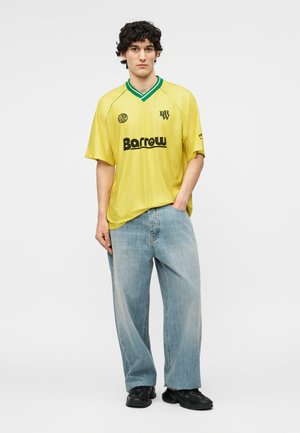 Ung mann med krøllete hår som har på seg gul kortermet skjorte med "Barrow"-logo, løse lyseblå jeans og svarte sko, står med den ene hånden i lommen.