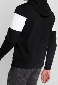 Felpa nera con pannelli bianchi sulle spalle, realizzata in tessuto strutturato e dotata di una tasca a marsupio, rifinita con polsini e orlo a coste.