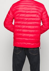 Röd vadderad jacka med ett quiltat mönster, dragkedjefickor på sidorna och en slät, glänsande yta. Buren över mörka denimbyxor.