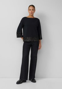 Schwarzes Oberteil mit dreiviertellangen Ärmeln und einem mit Pailletten verzierten Saum. Kombiniert mit dunklen, weit geschnittenen Jeanshosen und glänzenden schwarzen Schuhen.