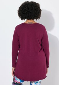Langärmlige Tunika in tiefem Lila mit abgerundetem Saum und glatter Textur. Kombiniert mit gemusterten Leggings in Blau- und Weißtönen.