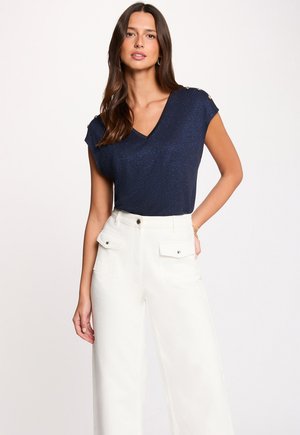 Donna con indosso una maglia blu navy a maniche corte con scollo a V e bottoni sulle spalle, e pantaloni a vita alta bianchi a gamba larga, che posa con la mano sul fianco.