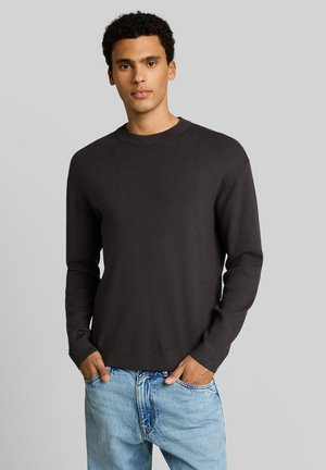 Jeune homme aux cheveux bouclés portant un pull noir à manches longues et un jean bleu clair, les mains dans les poches, debout devant un fond uni.