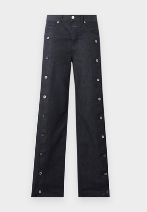 Persona che indossa occhiali da sole scuri, camicia oversize a quadri verdi e viola sopra un top rosa, pantaloni scuri con bottoni, scarpe marroni, che tiene una grande borsa marrone texturizzata.